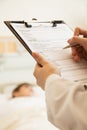 Close up of doctor writing on a medical chart with patient lying in a hospital bed in the background Royalty Free Stock Photo