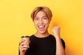 Close-up of energized handsome asian guy fist pump joyfully while drinking coffee, smiling excited over yellow