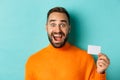 Close-up of excited caucasian man showing his credit card, smiling and staring amazed, standing in orange sweater Royalty Free Stock Photo