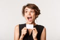 Close-up of excited young pretty woman, looking left ecstatic, showing credit card, standing over white background