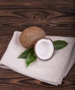 Close-up of an exotic whole and cracked in half coconuts with green leaves on a grey fabric and on a dark brown wooden background.