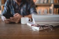 Close up eyeglasses and books stack on wooden desk in university or public library with man using mobile phone reading or text Royalty Free Stock Photo