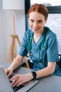 Close-up face of attractive smiling young female doctor in blue green medical uniform sitting at desk with laptop on Royalty Free Stock Photo