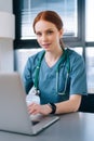 Close-up face of attractive smiling young female doctor in blue green medical uniform typing on laptop computer, sitting Royalty Free Stock Photo