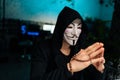 Close-up face of confident wanted hacker man wearing anonymous mask and hoodie holding beads in hands and looking away