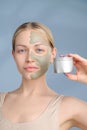 A close-up face portrait of a young blonde woman. Green beauty mask on half of the face. A woman holds a jar of Royalty Free Stock Photo