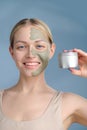 A close-up face portrait of a young blonde woman. Green beauty mask on half of the face. A woman holds a jar of Royalty Free Stock Photo