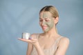 A close-up face portrait of a young blonde woman. Green beauty mask on half of the face. A woman holds a jar of Royalty Free Stock Photo