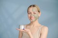 A close-up face portrait of a young blonde woman. Green beauty mask on half of the face. A woman holds a jar of Royalty Free Stock Photo