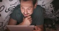 Close up of face of a young bearded and mustashed man, using digital tablet under blanket, in his bed, at night, and