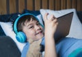 Close up face young boy wear wireless headphone listening music lying in bed,Happy Kid playing game on tablet, Smiling child in Royalty Free Stock Photo