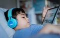 Close up face young boy wear wireless headphone listening music while sitting in bed,Cute kid playing game on tablet,Positive Royalty Free Stock Photo