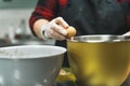 Close-up of female baker wearing gloves and black apron cracking egg into golden bowl to prepare cupcake batter