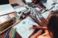 Close-up of female designer drawing floral compositions with crayons sitting at workplace surrounded with paint, gouache