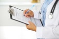 Close-up of a female doctor filling up medical form at clipboard while standing straight in hospital