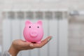 Close up of a female hand holding piggy bank with heating radiator on background Royalty Free Stock Photo