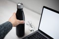 Close-up of female hand holding steel eco thermo bottle for water over office desk, laptop with mockup and glasses.