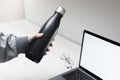 Close-up of female hand holding steel eco thermo bottle for water over office desk, laptop with mockup and glasses. Royalty Free Stock Photo