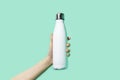 Close-up of female hand, holding white reusable steel stainless eco thermo water bottle on background of cyan, aqua menthe color.