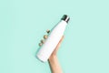 Close-up of female hand, holding white reusable steel stainless eco thermo water bottle with mockup, isolated on cyan color.