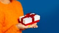 Close-up of female hands holding white gift box with red bow on background of blue color with copy space. Wearing orange sweater. Royalty Free Stock Photo