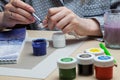 A close-up of female hands over a painted picture holding a brush and a cover for watercolors on which the colors are mixed next