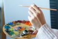 Close-up of female hands in a pink sweater holding a brush and a palette with multi-colored acrylic paints against a white canvas Royalty Free Stock Photo