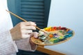 Close-up of female hands in a pink sweater holding a brush and a palette with multi-colored acrylic paints against a white canvas Royalty Free Stock Photo