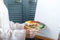 Close-up of female hands in a pink sweater holding a brush and a palette with multi-colored acrylic paints against a white canvas Royalty Free Stock Photo