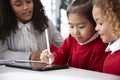 Close up of female infant school teacher sitting at a desk in a classroom helping two schoolgirls wearing school uniforms using a