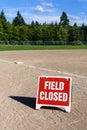 Close up of Field Closed sign on empty local baseball field, third base and baseline, on a sunny day with woods and blue sky in th