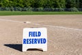 Close up of Field Reserved sign on empty local baseball field, third base and baseline, sunny day with woods in the background