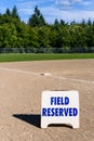 Close up of Field Reserved sign on empty local baseball field, third base and baseline, on a sunny day with woods and blue sky in Royalty Free Stock Photo