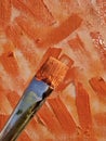 Oil paintbrush loaded with orange paint in front of scumbled canvas.