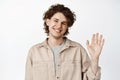 Close up of friendly smiling young man, waves his hand and smiles to greet you, say hello, stands over white background Royalty Free Stock Photo