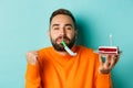 Close-up of funny adult man celebrating his birthday, holding bday cake with candle, blowing party wistle and rejoicing