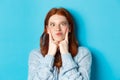 Close-up of funny redhead teen girl making faces, squinting and pocking cheeks, standing against blue background Royalty Free Stock Photo