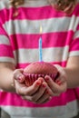 A girl in a pink and white shirt holding a pink cupcake in two hands with a blue burning candle, vertical Royalty Free Stock Photo