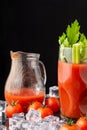 Close-up of glass with Bloody Mary cocktail with celery, jug with tomato juice, ice and cherry tomatoes, on black background, in v