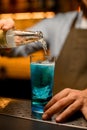 Close-up of glass of bright blue cocktail with piece of ice in which pouring liquid from bottle Royalty Free Stock Photo