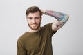 Close up of good-looking cool european guy with beard and arm tattoo smiling brightfully, holding hand behind head with Royalty Free Stock Photo