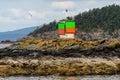 Close up of green and orange navigation marker on a rock in the Salish Sea, forested island and cloudy sky in background, San Juan