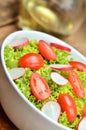 Close-up of green salad Lollo Biondo with tomatoes and radishes in a white bowl on wooden table, a pitcher of oil in the