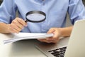 Close up hand of businessperson looking at documents through magnifying glass and laptop in front.