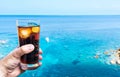 Hand holding drinking glass with ice-cooled cola against blue sky and sea Royalty Free Stock Photo