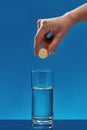 Close Up Of Hand Holding Effervescent Aspirin Tablet, Pill Above Glass Of Water Isolated Over Blue Background. Health