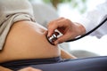 Hand of male obstetrician doctor with stethoscope listening to pregnant woman baby heartbeat.