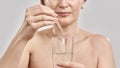 Close up of hand of middle aged woman holding dropper with medication and glass isolated over grey background