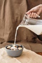 Close up of hand pouring, adding milk to homemade granola in a plate with nuts, honey, berries and other natural