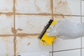 Close-up of a hand in a protective glove, holding a yellow spatula, in the process of grout white tiles, light brown sealant Royalty Free Stock Photo